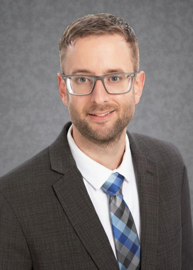 Headshot of Marcus Tabert, wearing glasses, a dark suit, and a blue plaid tie against a gray studio background.