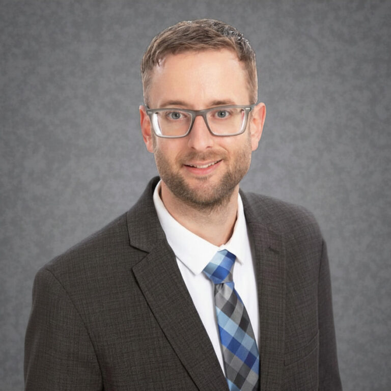 Headshot of Marcus Tabert, wearing glasses, a dark suit, and a blue plaid tie against a gray studio background.