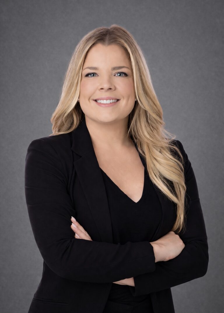 Headshot of Megan Coleman, smiling with arms crossed in a black blazer against a gray studio background.