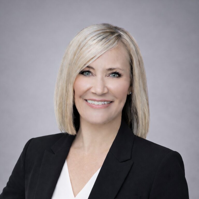Headshot of Tami Greene, smiling with shoulder-length blonde hair, wearing a black blazer over a white blouse.