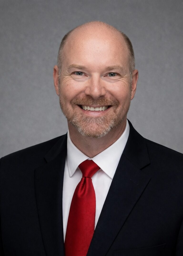 Headshot of Trent Borts, smiling in a dark suit and red tie against a gray background.