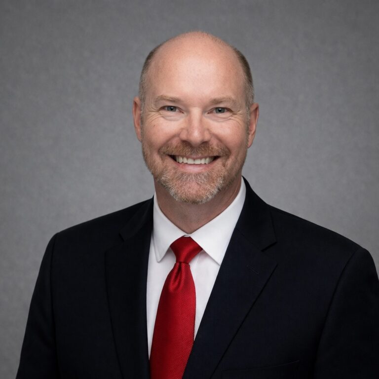 Headshot of Trent Borts, smiling in a dark suit and red tie against a gray background.