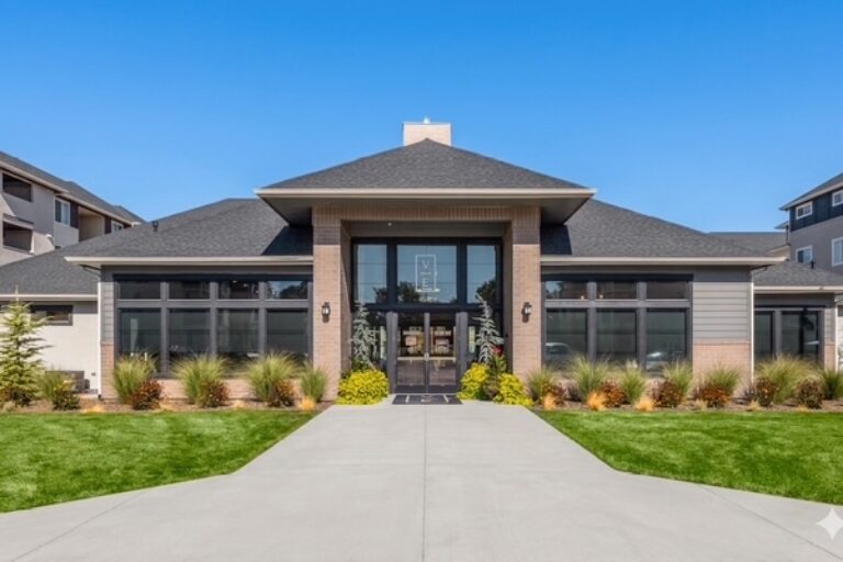 Modern apartment clubhouse with a wide glass entrance, landscaped shrubs, and a concrete walkway under a clear blue sky