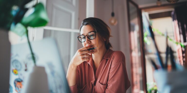 Woman with glasses thinking at a desk, holding a pen while looking at a laptop in a softly lit home office