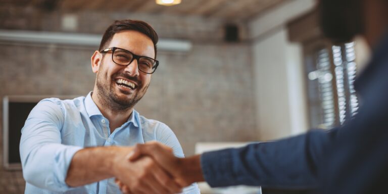 Smiling man in glasses shaking hands with another person across a table in an office.