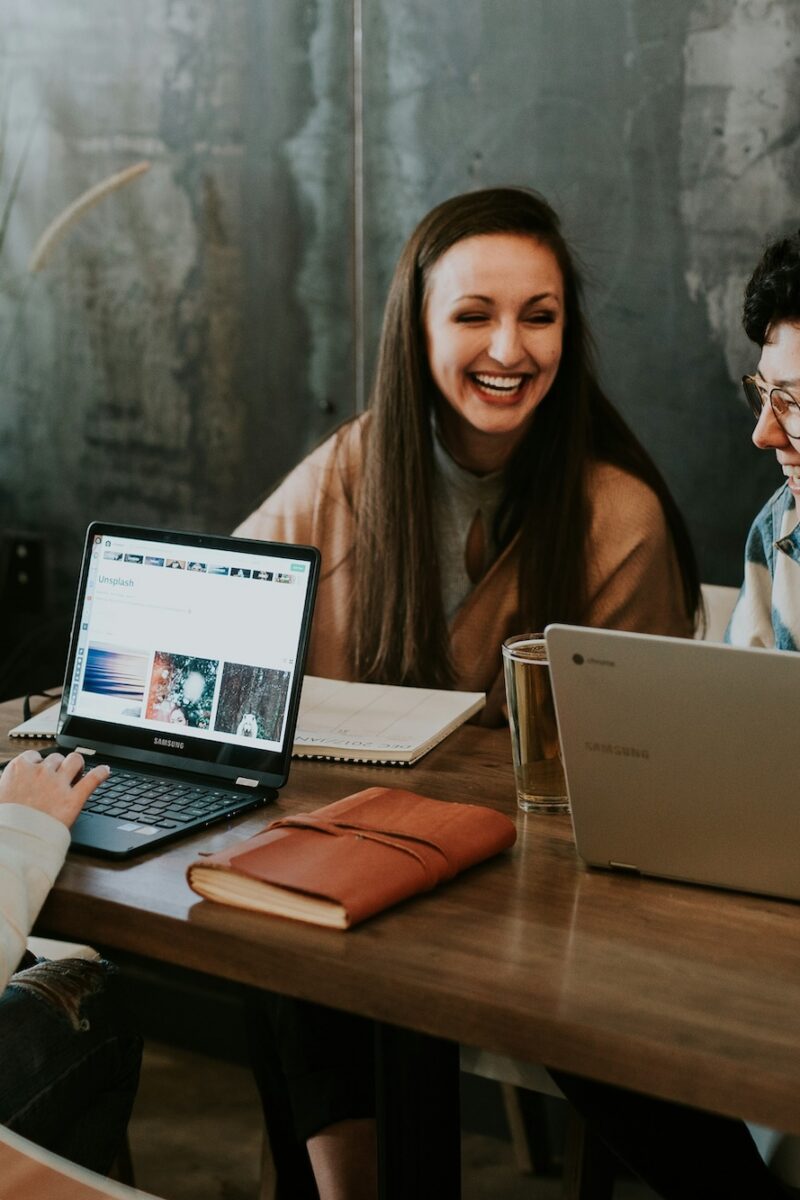 Three people laughing together at a table with laptops, drinks, and a notebook in a casual meeting space