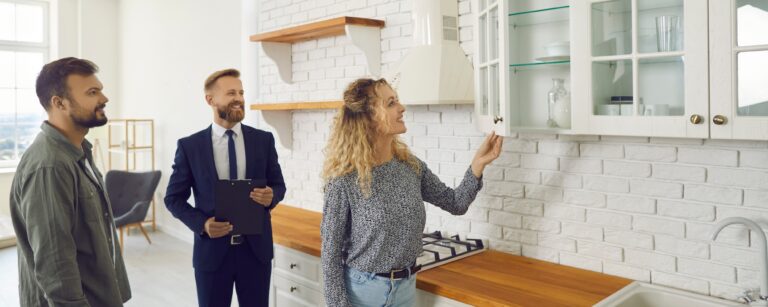 Couple touring a bright white kitchen with a real estate agent as the woman opens a glass-front cabinet