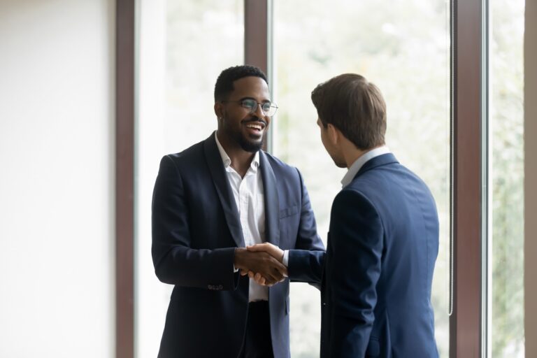 Two businessmen in suits smiling and shaking hands by a large office window