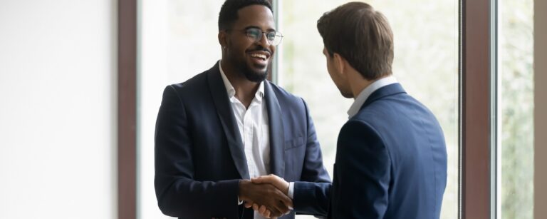 Two businessmen in suits smiling and shaking hands by a large office window