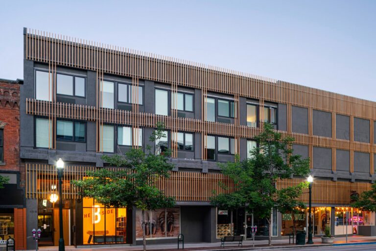 Modern three-story building with dark facade, vertical wood slats, and street-level storefronts along a tree-lined street