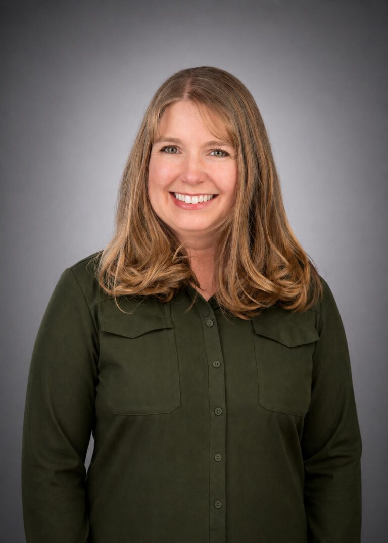 Smiling woman with shoulder-length light brown hair in a dark green button-up shirt against a gray studio background