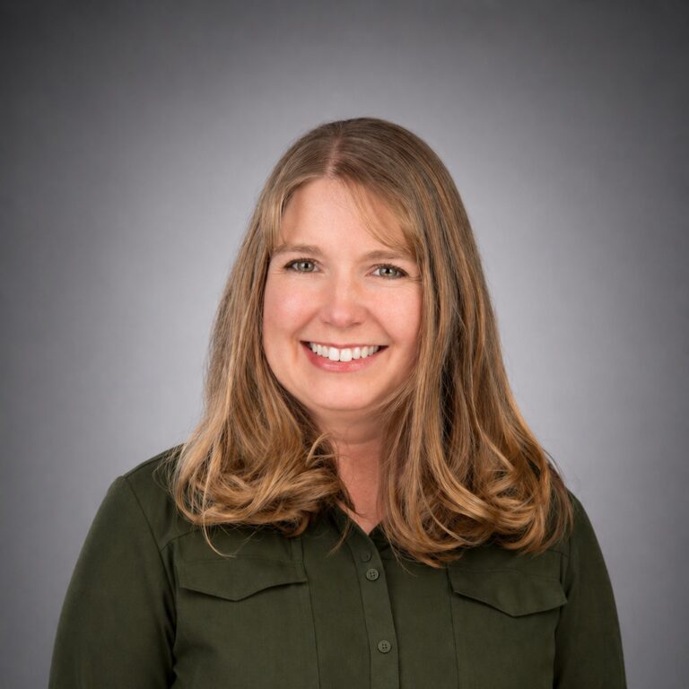 Smiling woman with shoulder-length light brown hair in a dark green button-up shirt against a gray studio background