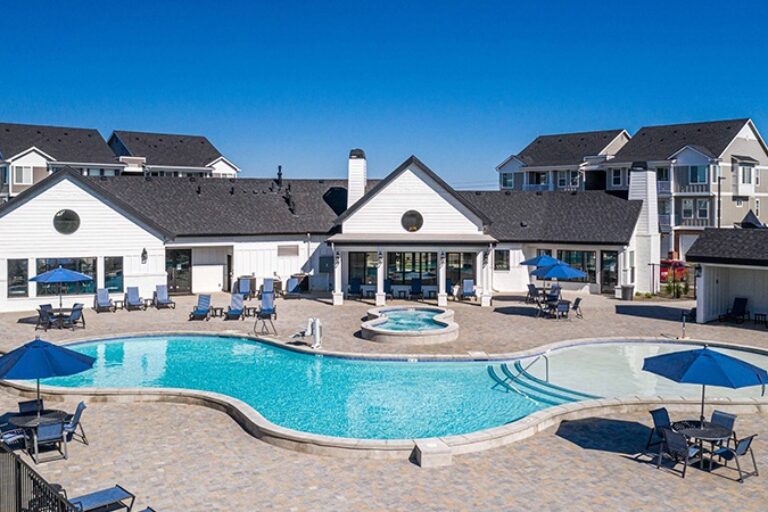Outdoor community pool with lounge chairs and blue umbrellas in front of a white clubhouse and apartment buildings