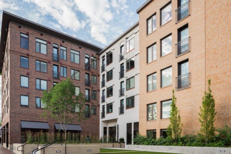 Modern apartment courtyard framed by brick and white mid-rise buildings with large windows, trees, and a small lawn