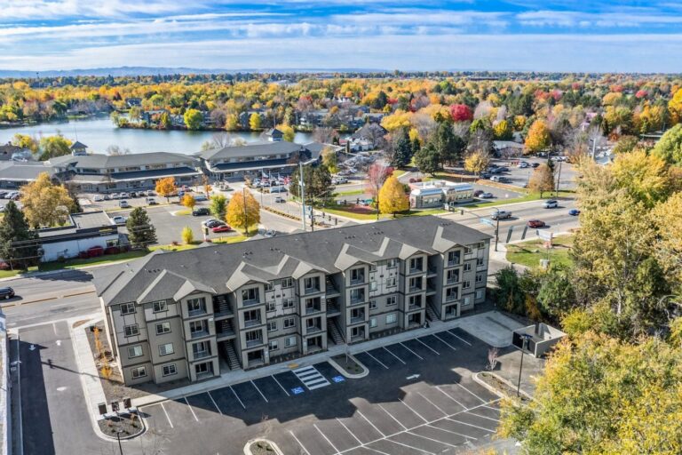 Aerial view of a four-story apartment building with parking lot, near a lake and colorful autumn trees.