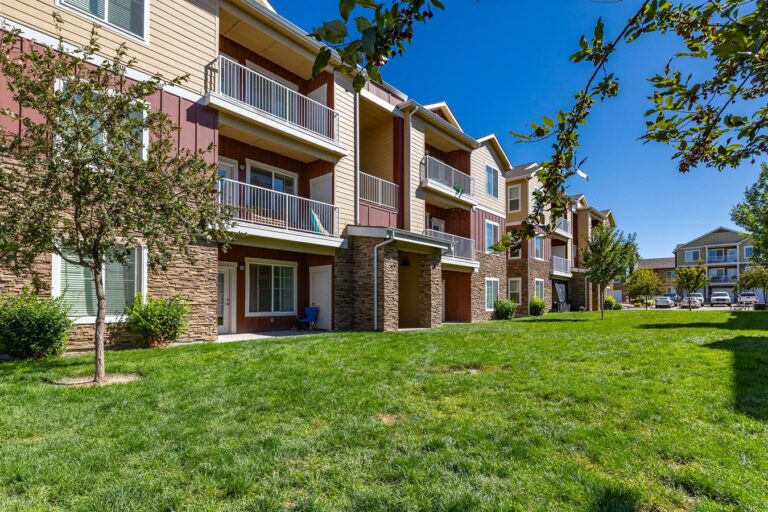 Three-story apartment buildings with balconies facing a grassy courtyard with trees under a clear blue sky