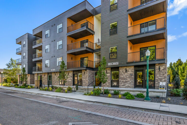 Modern four-story apartment building with brick base, dark siding, and stacked balconies along a quiet street.