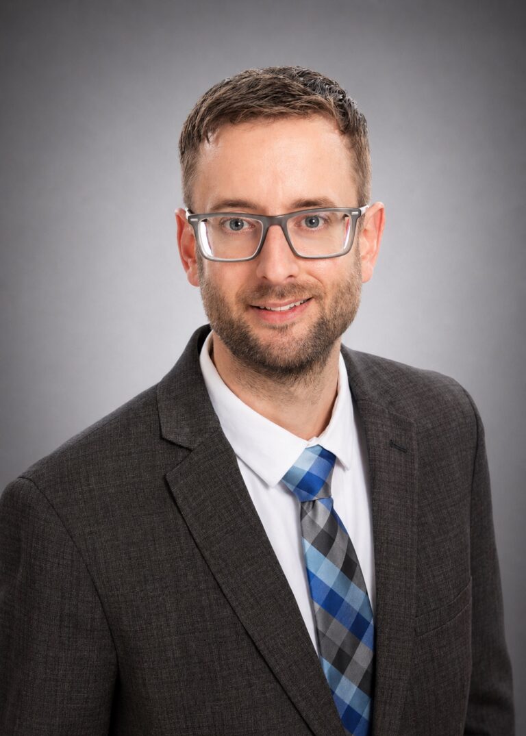 Professional headshot of a smiling man wearing glasses, a dark suit, and a blue plaid tie against a gray background
