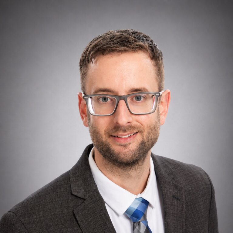 Professional headshot of a smiling man wearing glasses, a dark suit, and a blue plaid tie against a gray background
