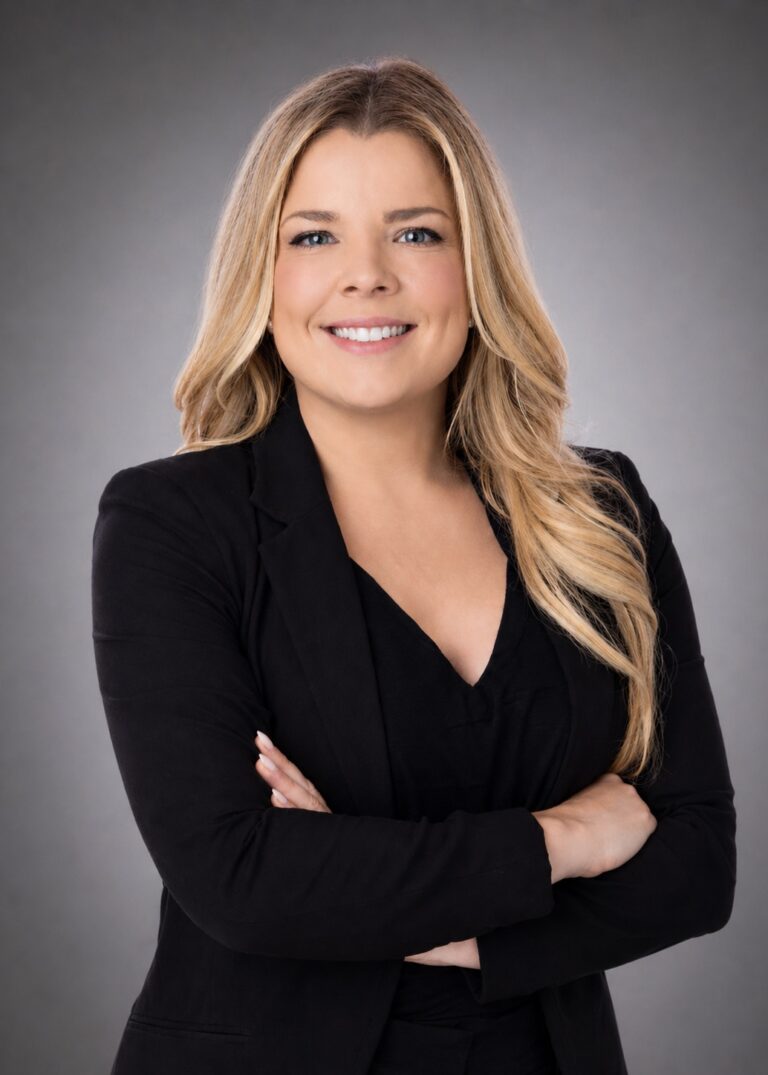 Smiling woman with long blonde hair in a black blazer, arms crossed, against a gray studio background