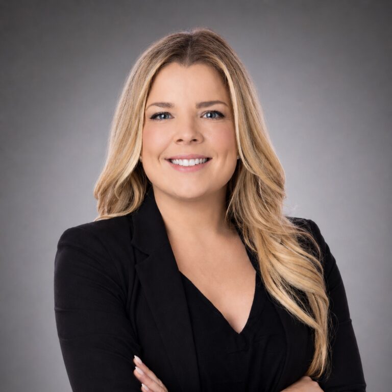 Smiling woman with long blonde hair in a black blazer, arms crossed, against a gray studio background