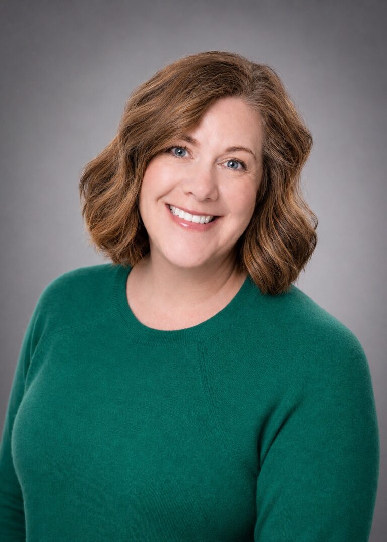 Smiling woman with shoulder-length wavy brown hair in a green sweater against a gray studio background