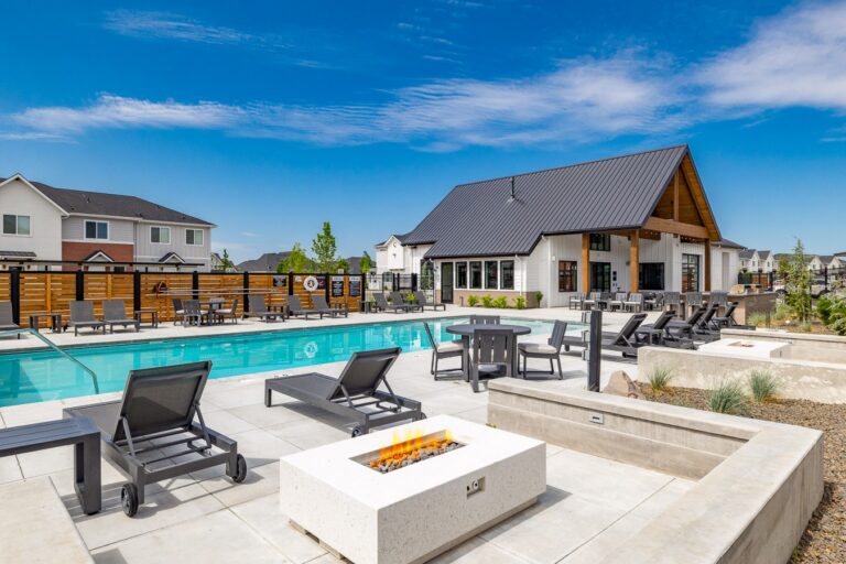 Modern outdoor pool area with lounge chairs, fire pits, and a clubhouse beside townhomes under a bright blue sky