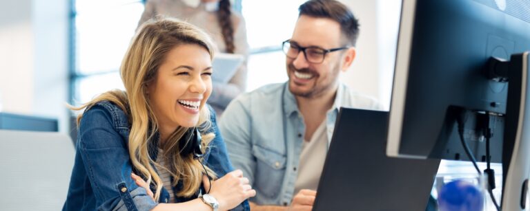 Three coworkers smiling and laughing together at a computer in a bright office.