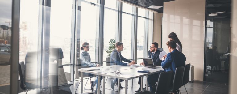 Five business professionals meeting around a table in a bright glass-walled office conference room