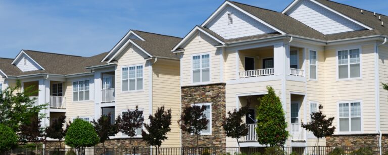 Three-story apartment building with beige siding, stone accents, balconies, and trees along the street