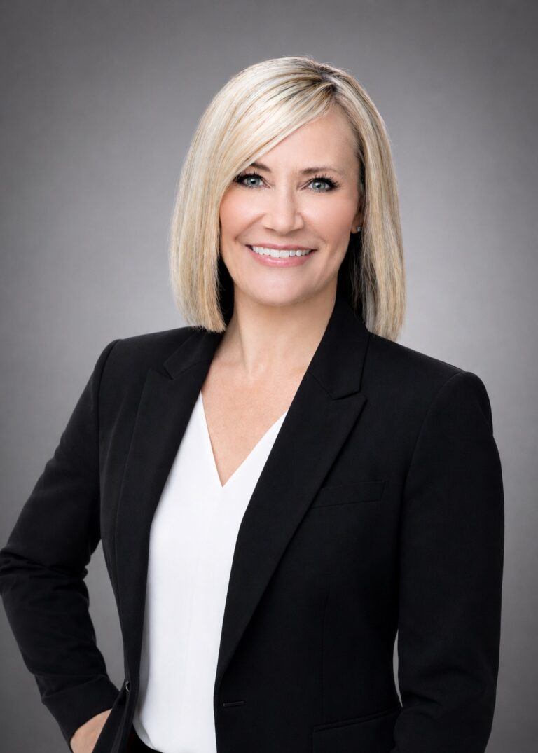 Professional headshot of a smiling blonde woman in a black blazer and white blouse against a gray studio background