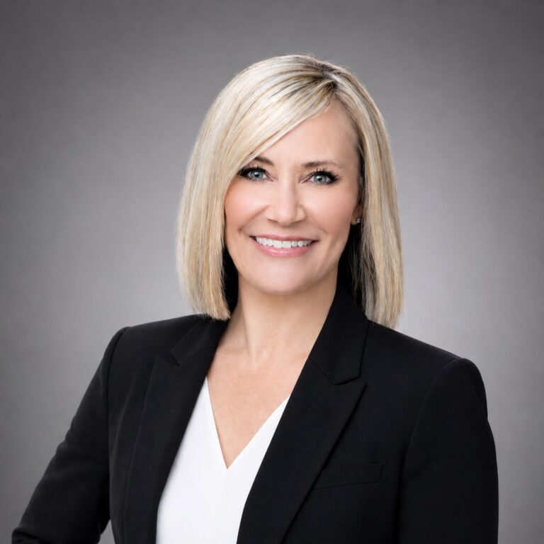 Professional headshot of a smiling blonde woman in a black blazer and white blouse against a gray studio background