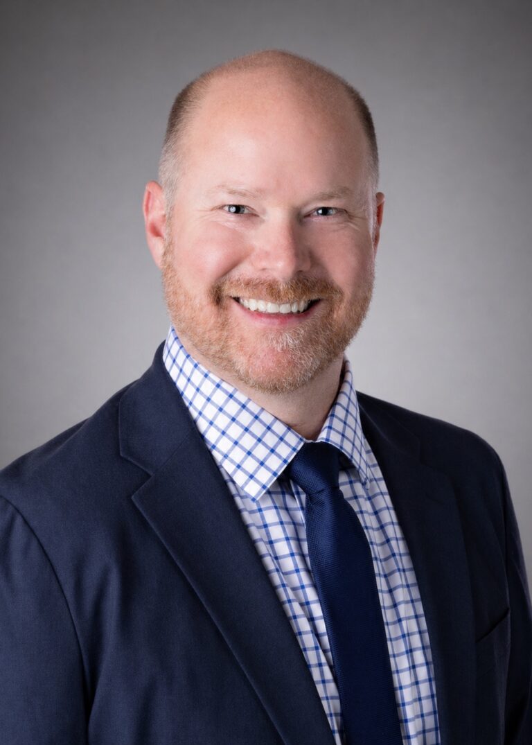 Smiling bald man with a short beard in a navy suit, checkered shirt, and tie against a gray studio background
