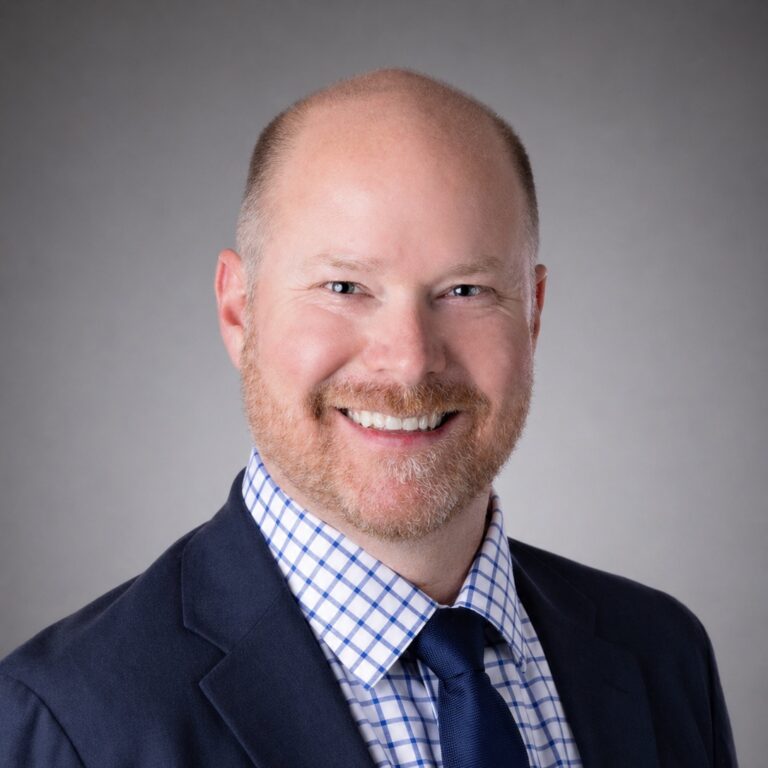 Smiling bald man with a short beard in a navy suit, checkered shirt, and tie against a gray studio background