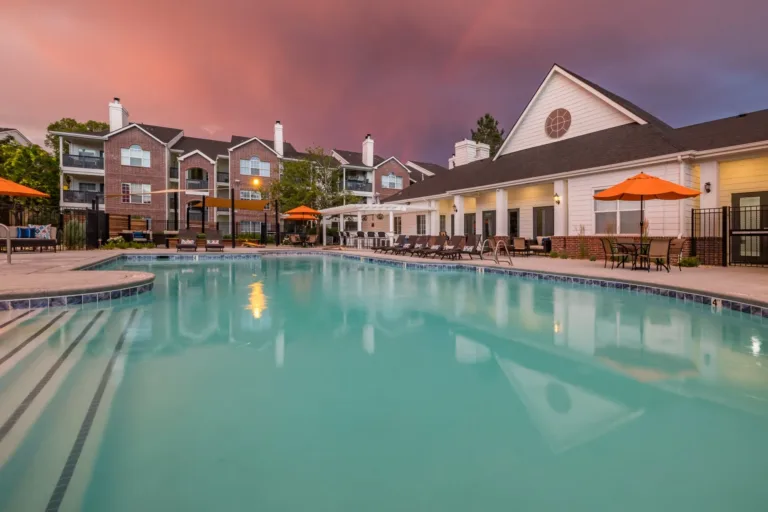 Apartment complex pool with lounge chairs and orange umbrellas at dusk under a pink and purple sky